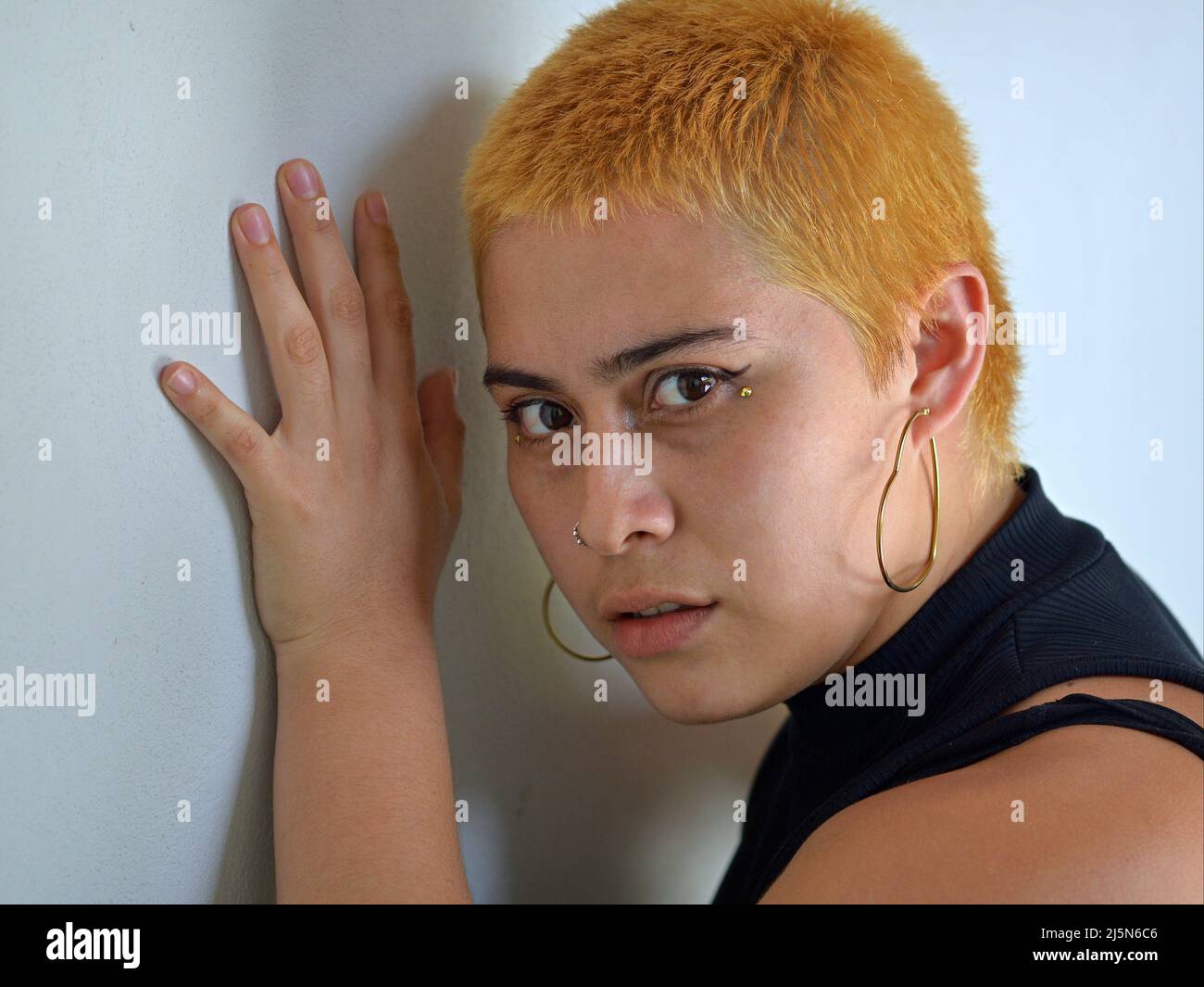 Giovane attraente alternativa messicana latina hipster donna con capelli corti tinti arancione e guarda lateralmente curiosamente al visore di fronte a una parete grigia. Foto Stock