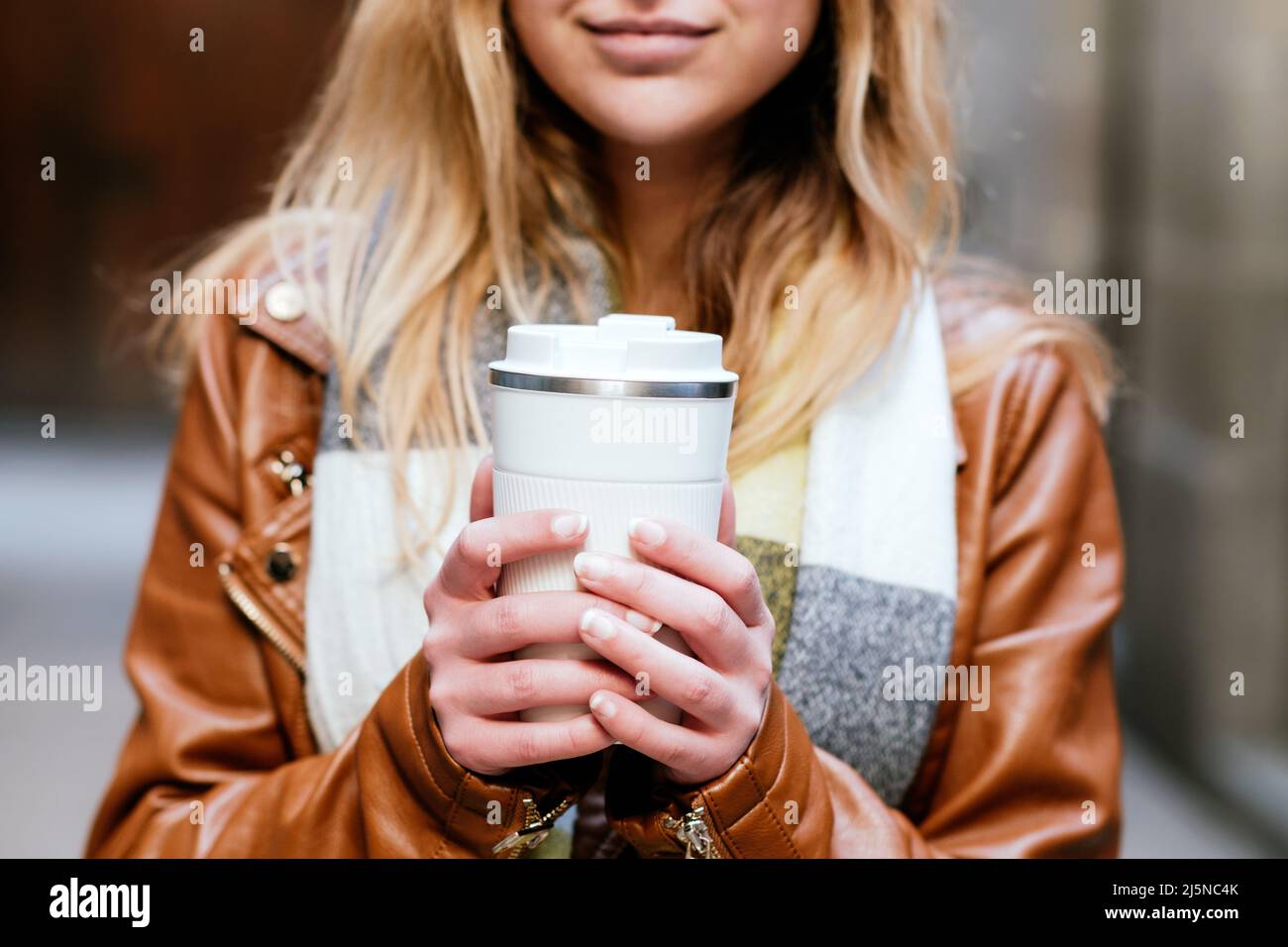 Crop anonymous femminile in giacca di pelle che tiene thermo tazza di bevanda calda mentre si trova sulla strada contro sfondo sfocato in città Foto Stock
