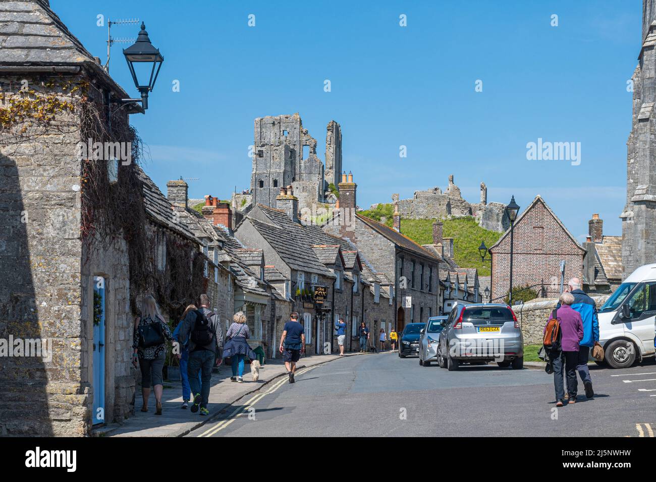 Corfe Castle Village Center, Dorset, Inghilterra, Regno Unito. Vista sulla strada del villaggio e del castello di Dorset, Inghilterra, Regno Unito, con i visitatori in una giornata di sole Foto Stock