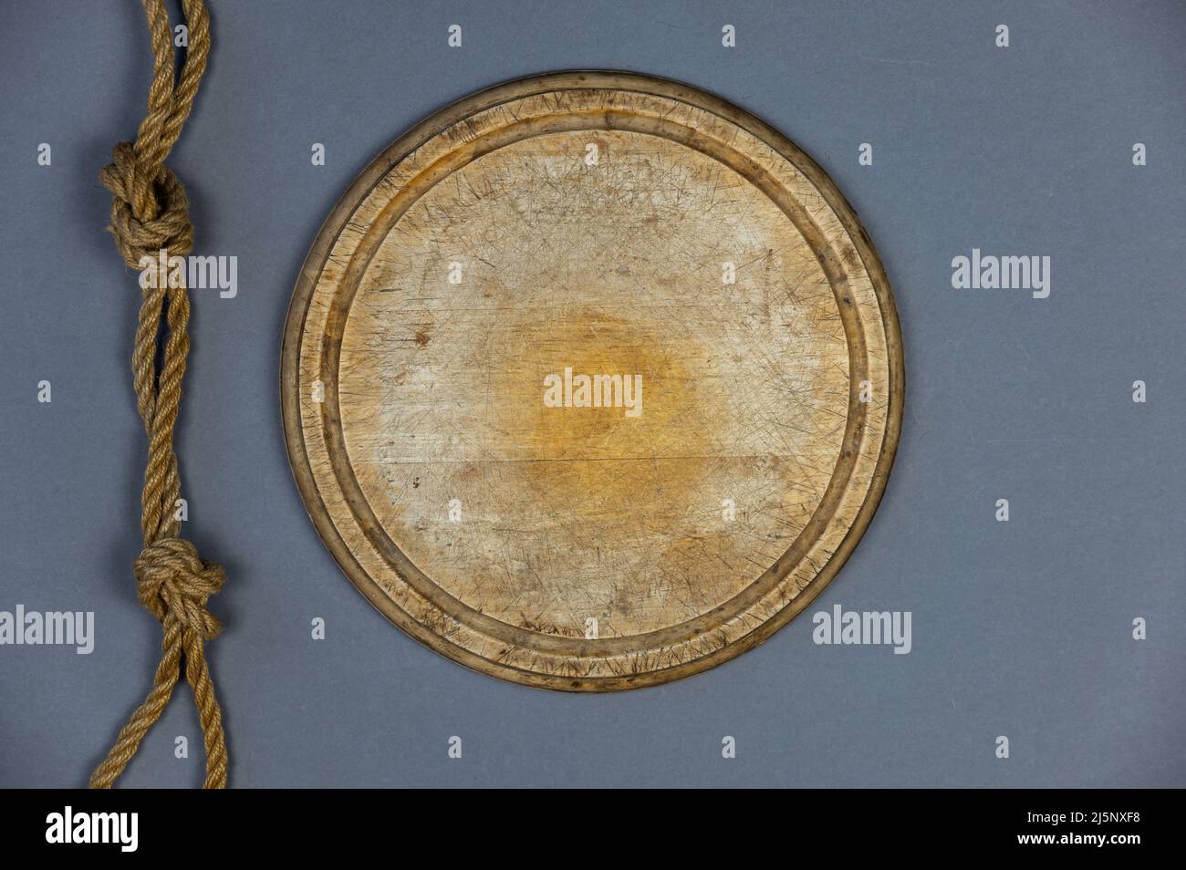 Tavola rotonda di taglio in legno e corda della nave sullo sfondo grigio. Svuotare il vecchio tagliere. Corda doppia di iuta con nodi semplici legati. Vista dall'alto. Foto Stock