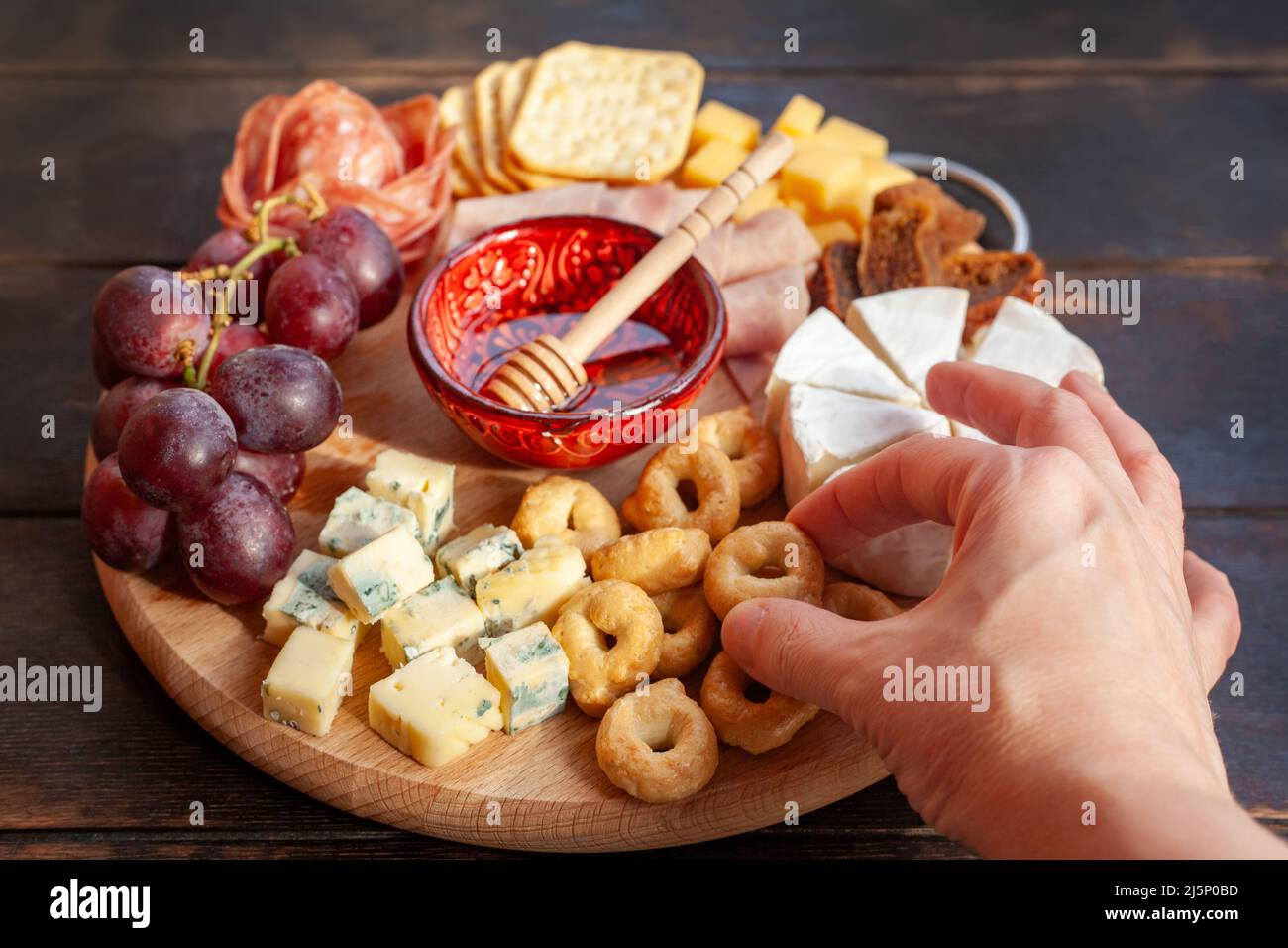La mano della donna prende un piccolo biscotto salato dal bordo degli antipasti con formaggio assortito, carne, rosetta di salsiccia, uva e biscotti. Salumi e chee Foto Stock