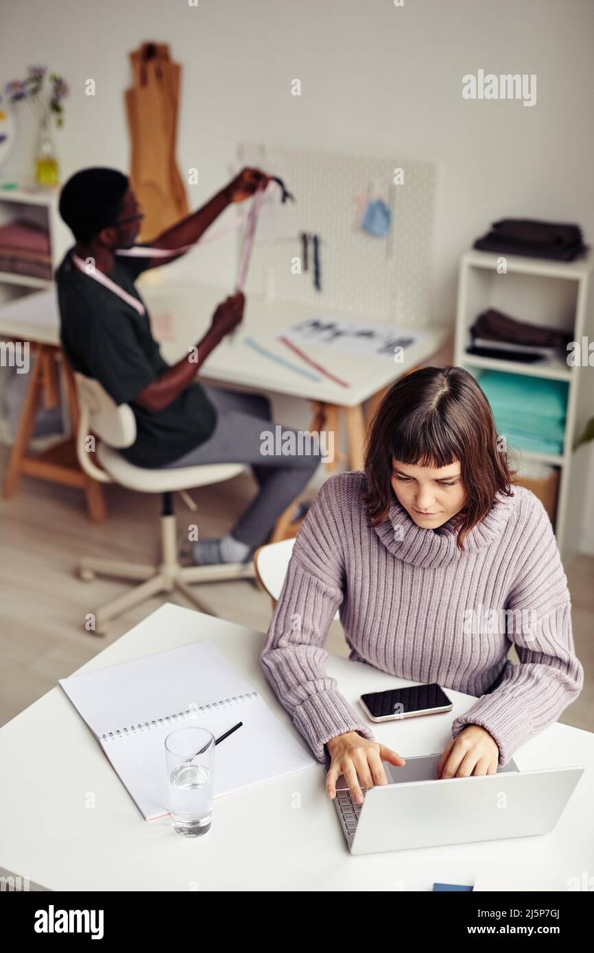 Giovane donna creativa che fa modelli di cucito su laptop quando stilista di moda lavorando su una nuova collezione in background Foto Stock