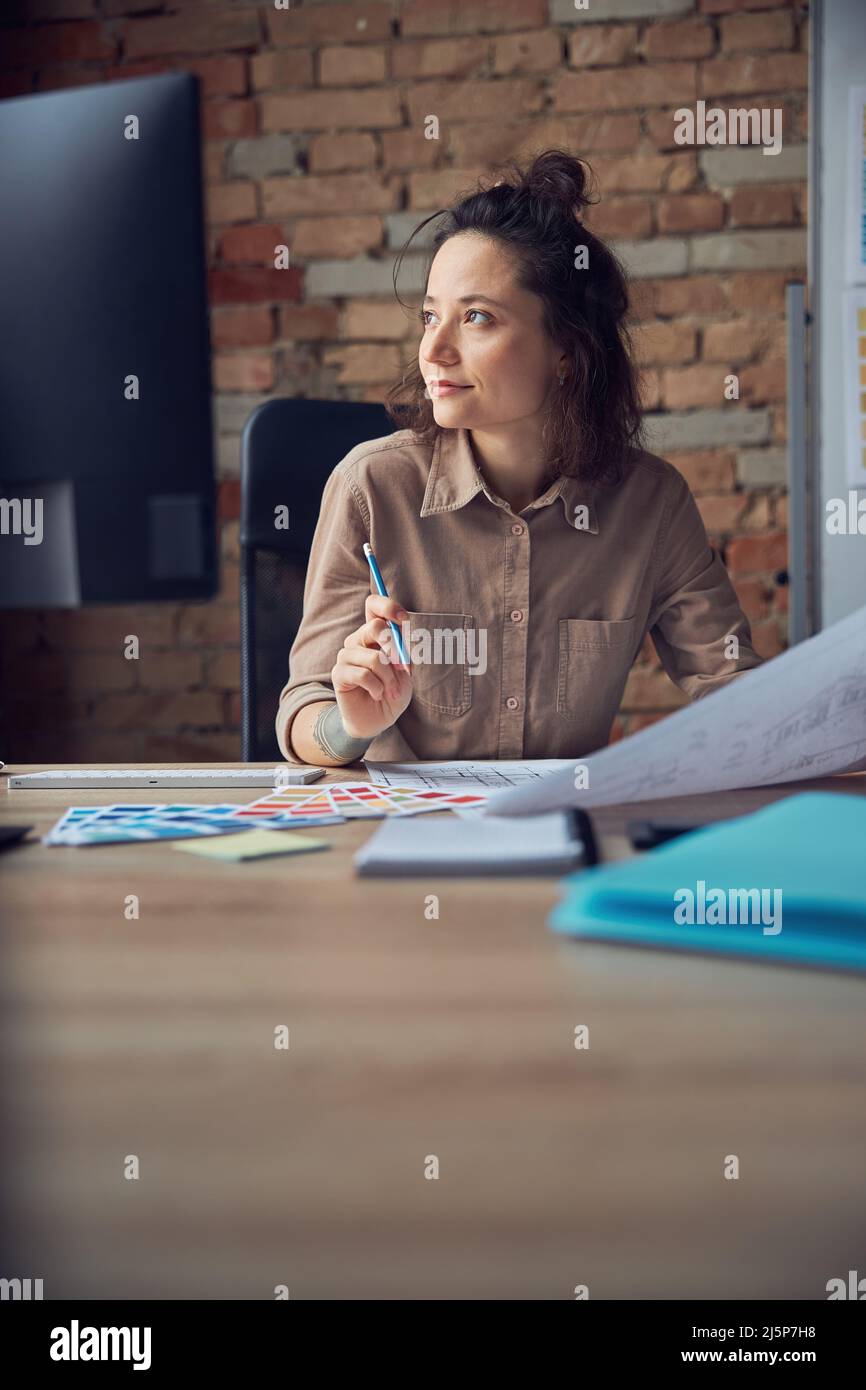 Allegra designer di interni femmina sorridente mentre lavora con disegno, campione di materiale nel suo ufficio Foto Stock