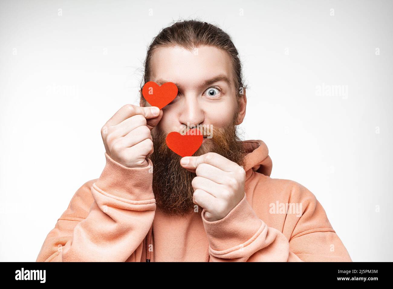 Ritratto di felice bell'uomo con acconciatura zenzero e barba su sfondo grigio. Attraente uomo scandinavo che è giocoso e tenendo cuori rossi Foto Stock
