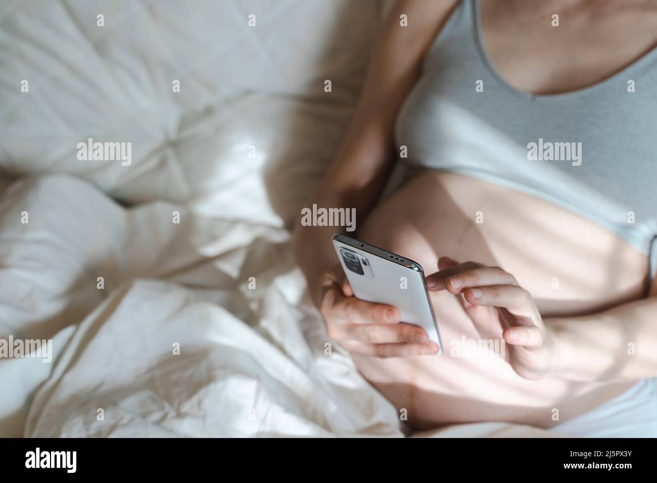 Una donna incinta usa uno smartphone mentre si rilassa nel suo letto di casa. Internet, shopping, comunicazione durante la gravidanza e prima della nascita di un bambino. Salute delle donne e dei neonati. Foto di alta qualità Foto Stock