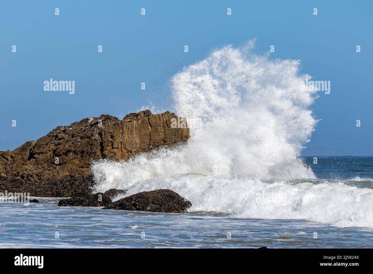 Onda gigante che si infrangono contro la grande roccia appena al largo di Malibu, California. Spruzzi che volano in alto nell'aria; cielo blu in lontananza. Foto Stock