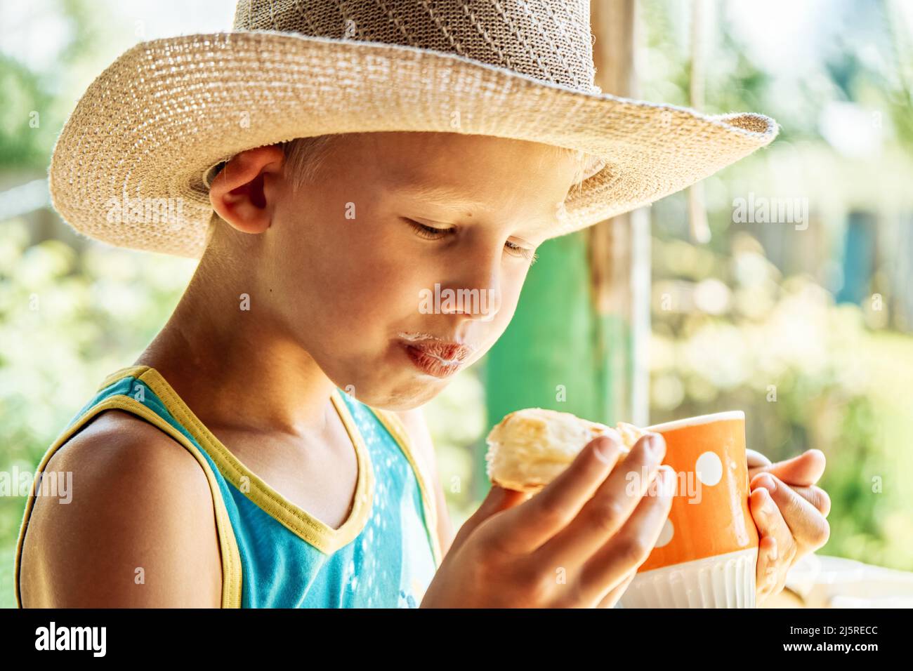 Il ragazzo di scuola carino in cappello di paglia ama bere il latte con la pistola sul giardino sfondo sfocato. Il bambino fa colazione all'aperto nel soleggiato primo piano del mattino Foto Stock