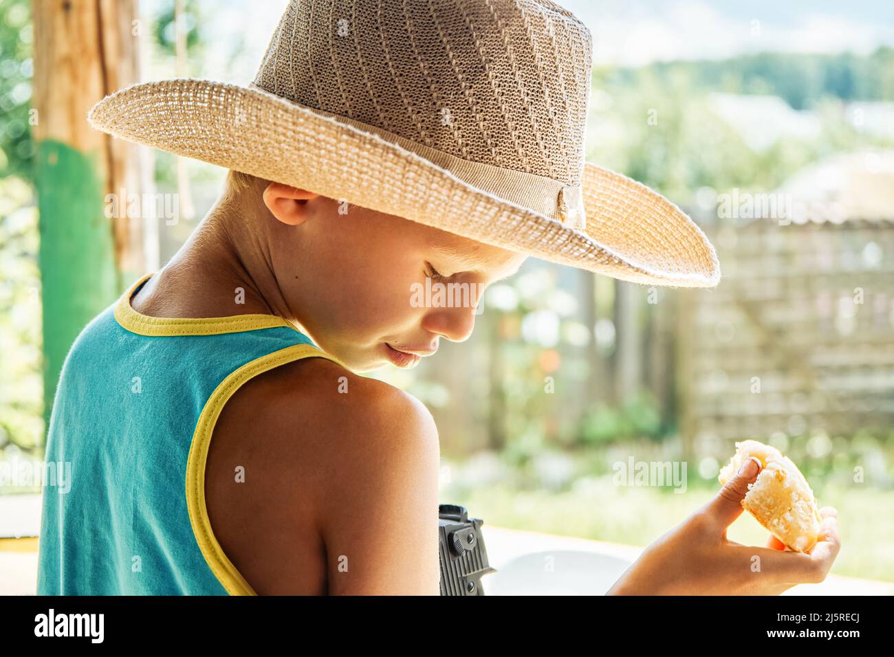 Ragazzo in paglia cappello mangia fresco pannolino in piedi sul portico in mattinata di sole. Carino schoolboy gode la prima colazione sulla casa cottage sfocato sfondo primo piano Foto Stock