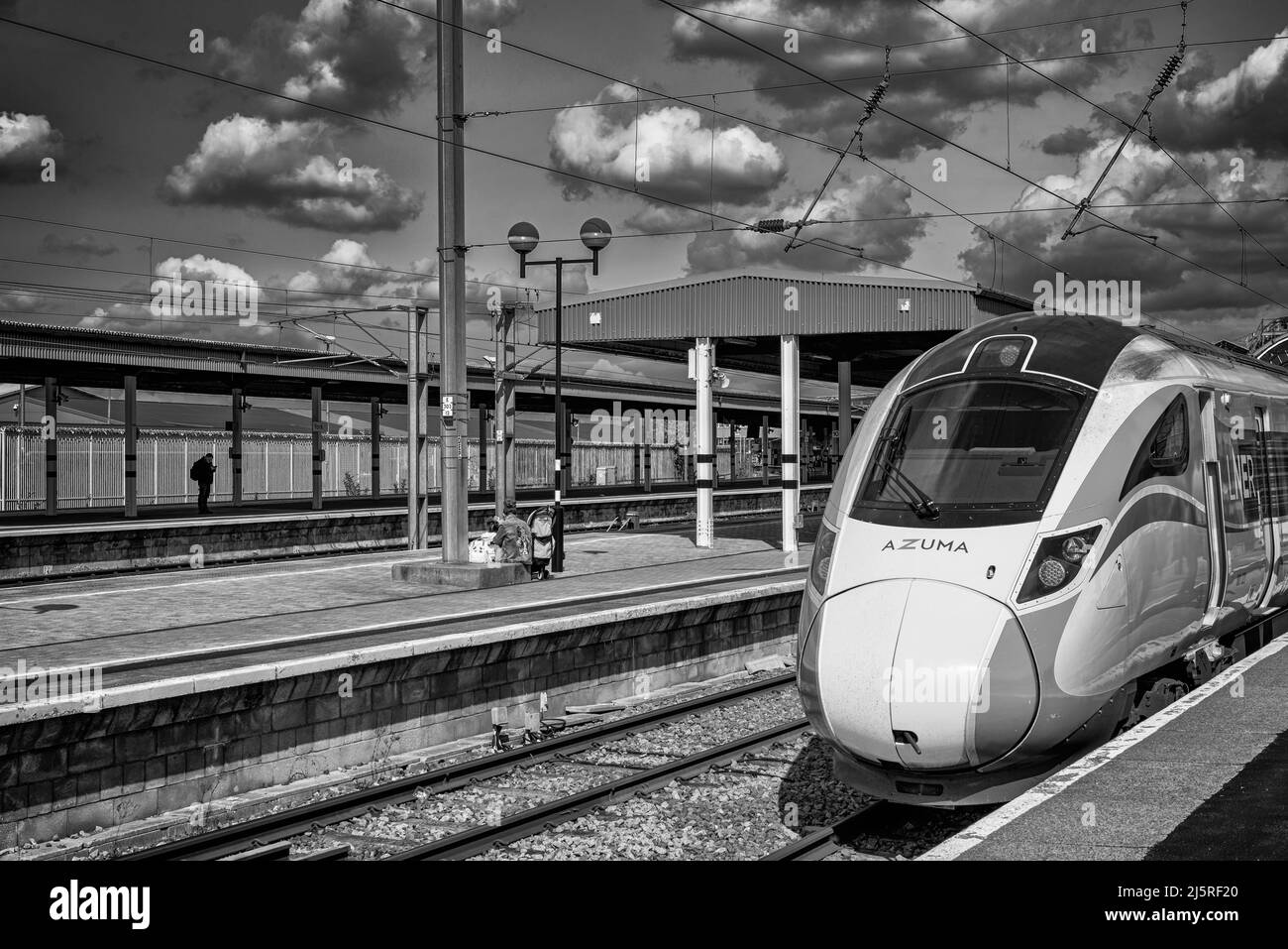 Un treno si trova su una piattaforma di una stazione ferroviaria. Una donna si siede con un bambino su una base di pietra e un cielo con la nuvola è sopra. Foto Stock