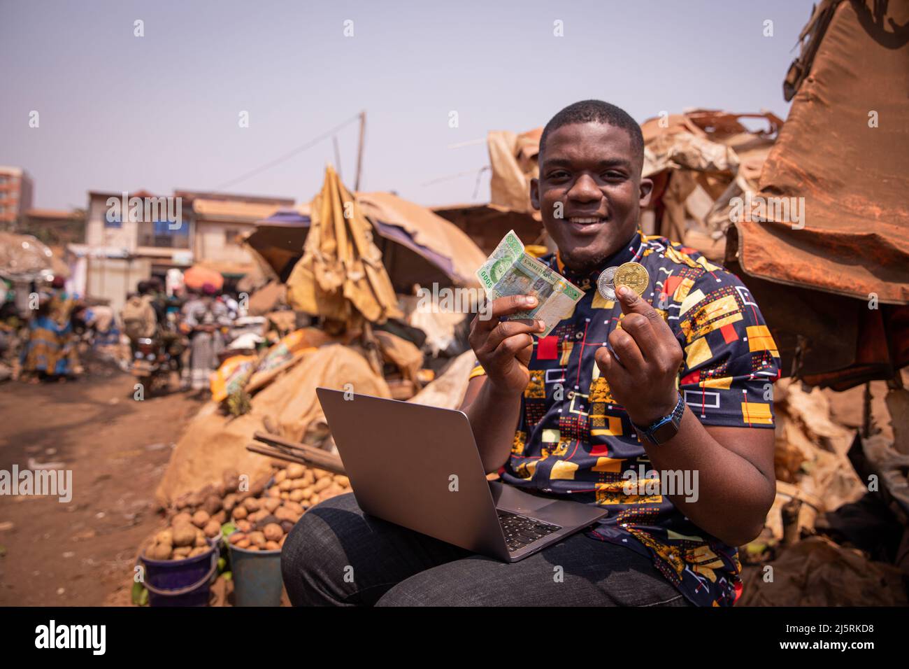 Ragazzo africano al mercato che detiene FCFA franchi e una moneta Bitcoin, l'attuazione delle criptovalute in africa Foto Stock