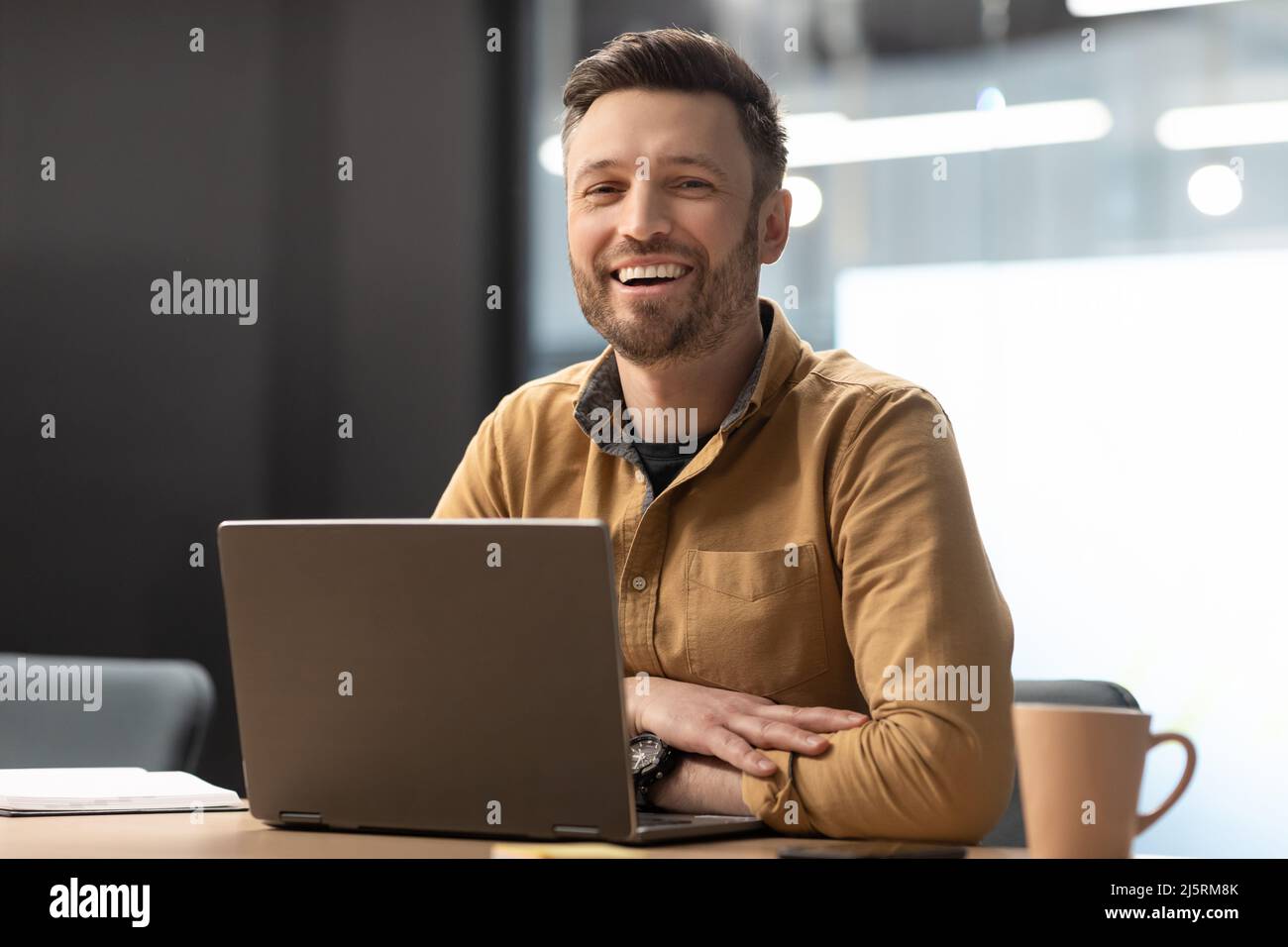 Imprenditore allegro maschio che posa vicino laptop che lavora in ufficio moderno Foto Stock