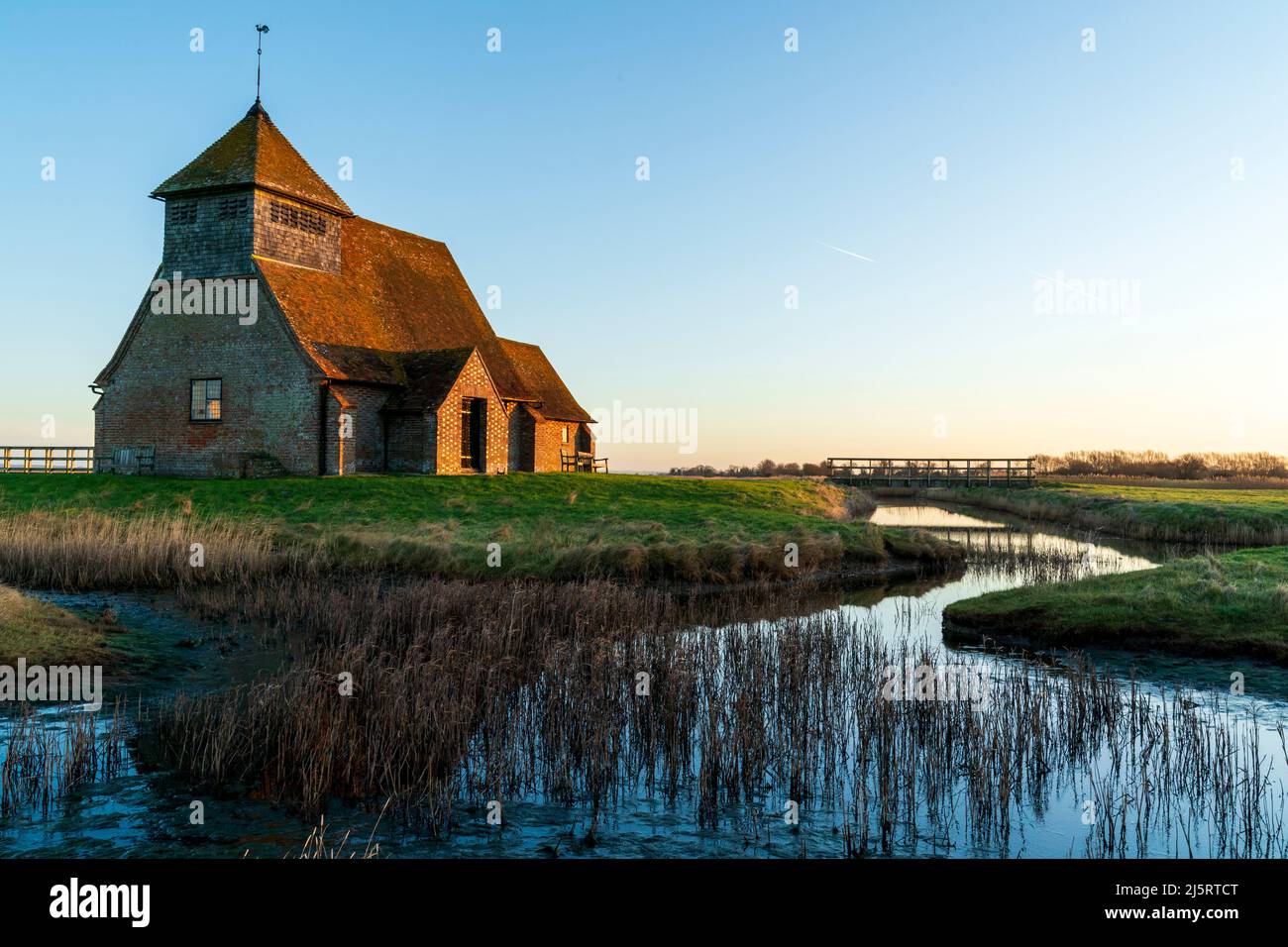 13th secolo chiesa di San Tommaso a Becket su Romney Marsh illuminato dalla luce dell'ora d'oro contro un cielo blu chiaro. Il primo piano è un flusso con Reeds. Foto Stock
