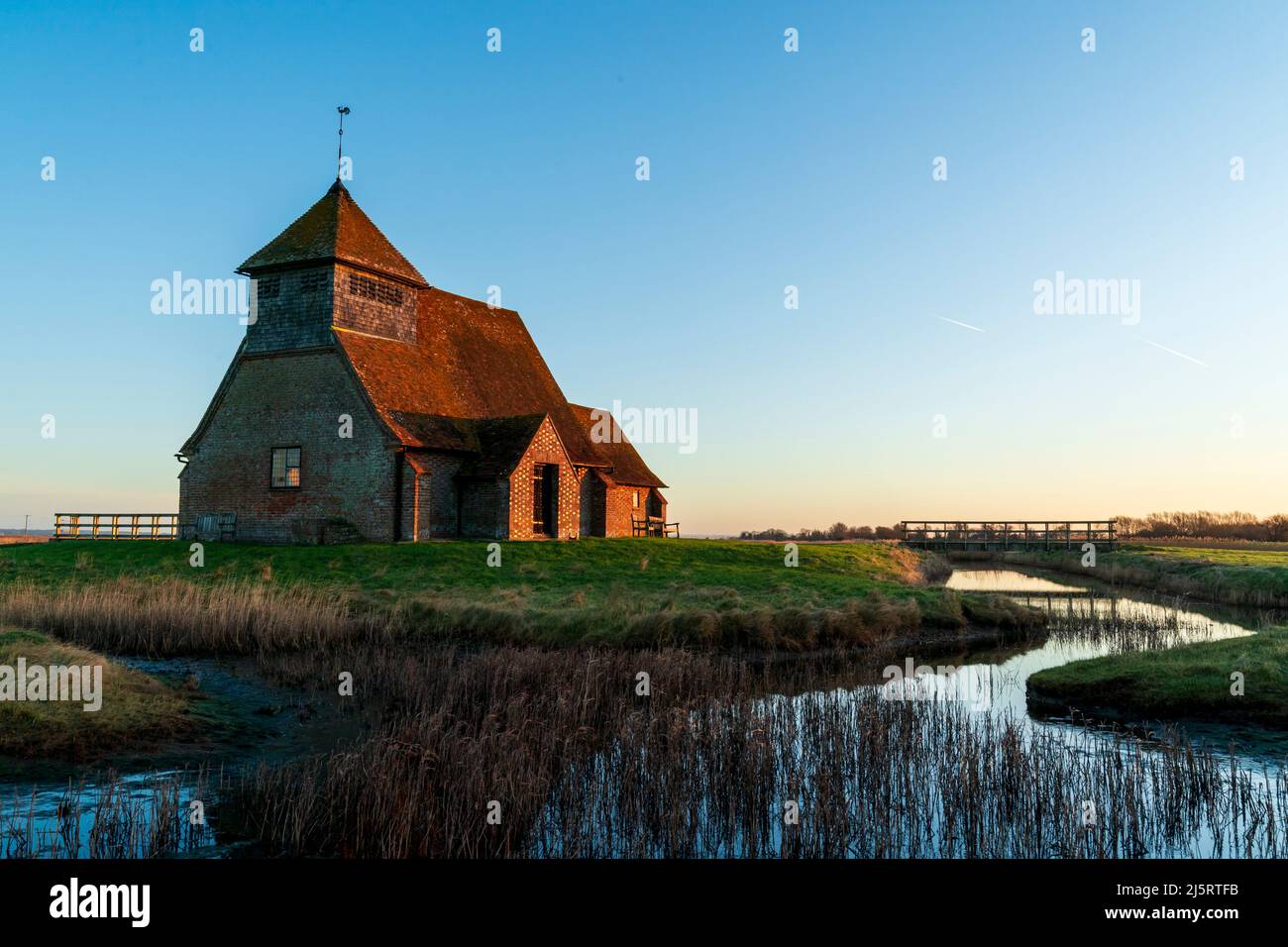 13th secolo chiesa di San Tommaso a Becket su Romney Marsh illuminato dalla luce dell'ora d'oro contro un cielo blu chiaro. Il primo piano è un flusso con Reeds. Foto Stock