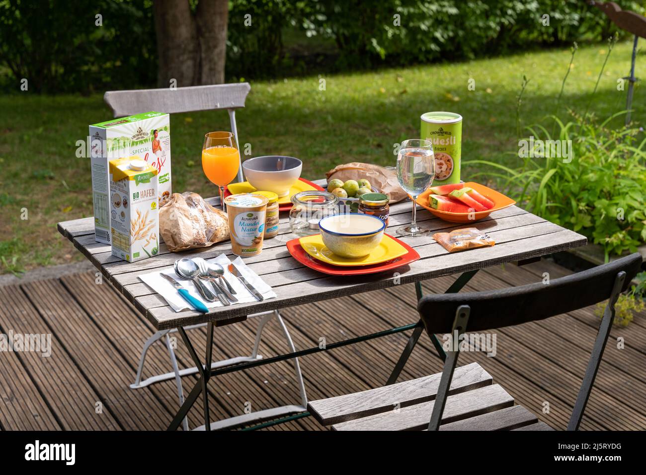 Prima colazione su un tavolo da giardino in legno. Cibo e bevande al mattino. Cereali, pane, latte e frutta. Prodotti diversi per un pasto sano. Foto Stock