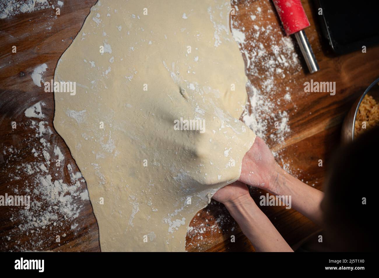 Vista dall'alto delle mani femminili che tirano e allungano un impasto di pasta vegan fatto in casa per strudel o torta su un tavolo da pranzo domestico. Foto Stock