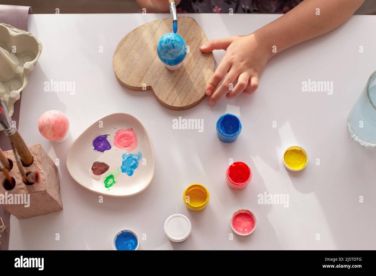 Le mani dei bambini dipingono un uovo, su un tavolo bianco con vernici multicolore in vasetti Foto Stock