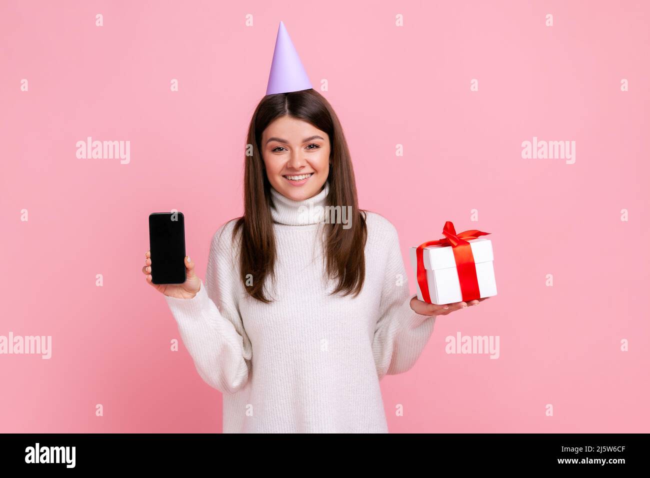 Donna ottimista felice in confezione regalo e smart phone con contenitore vuoto per l'annuncio, indossando un pullover casual bianco. Studio interno girato isolato su sfondo rosa. Foto Stock