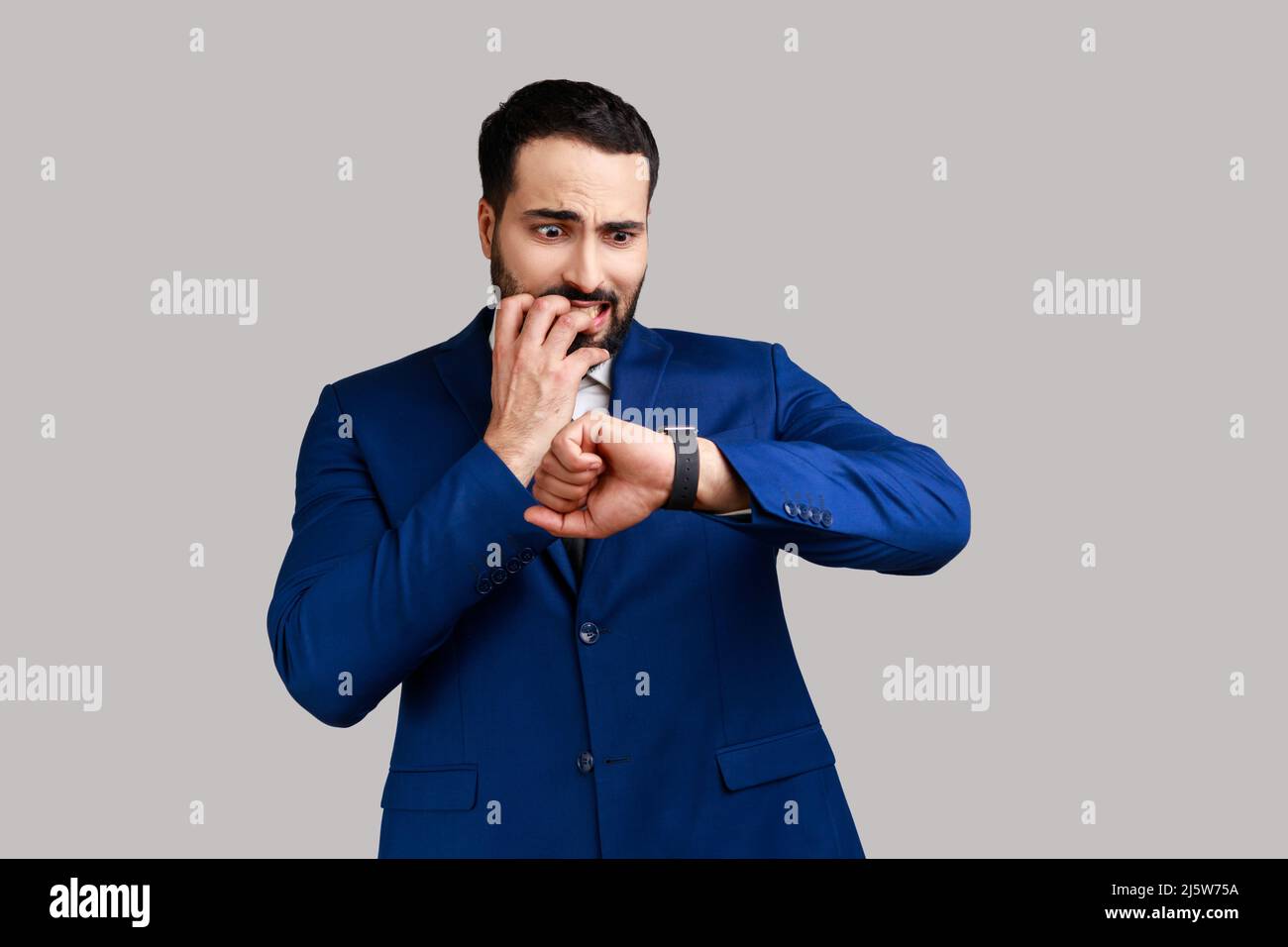L'uomo nervoso bearded che morde le unghie del dito e guarda il suo orologio del polso con ansioso, preoccupato circa la scadenza, che indossa il vestito ufficiale di stile. Studio interno girato isolato su sfondo grigio. Foto Stock