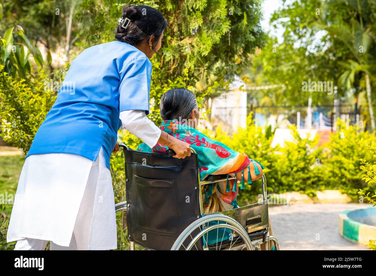 Back view shot of Nurse che prende la donna anziana ferita sulla sedia a rotelle a piedi al giardino dell'ospedale - concetto di custode, trattamento e assistenza sanitaria. Foto Stock