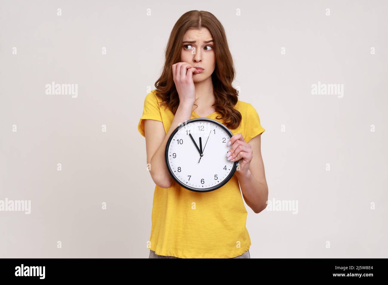 Impaziente adolescente nervoso ragazza con capelli ondulati in giallo t-shirt seduta mordente le sue unghie tenendo grande orologio da parete, scadenza, afrids per essere in ritardo. Studio interno girato isolato su sfondo grigio. Foto Stock