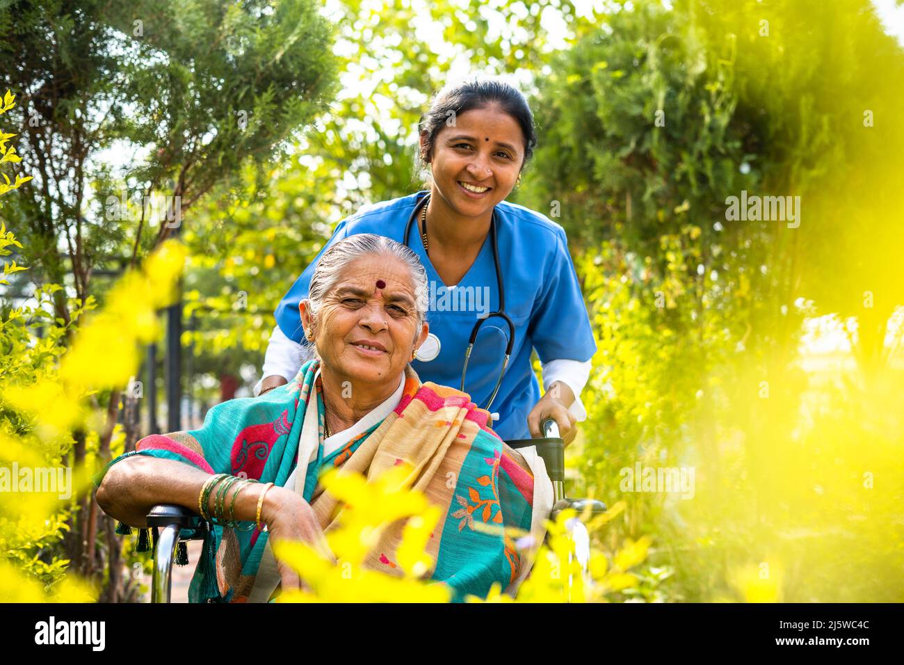 Infermiera sorridente felice che prende la donna anziana a piedi mentre sulla sedia a rotelle al giardino dell'ospedale guardando la macchina fotografica - concetto di custode, invalidità e. Foto Stock
