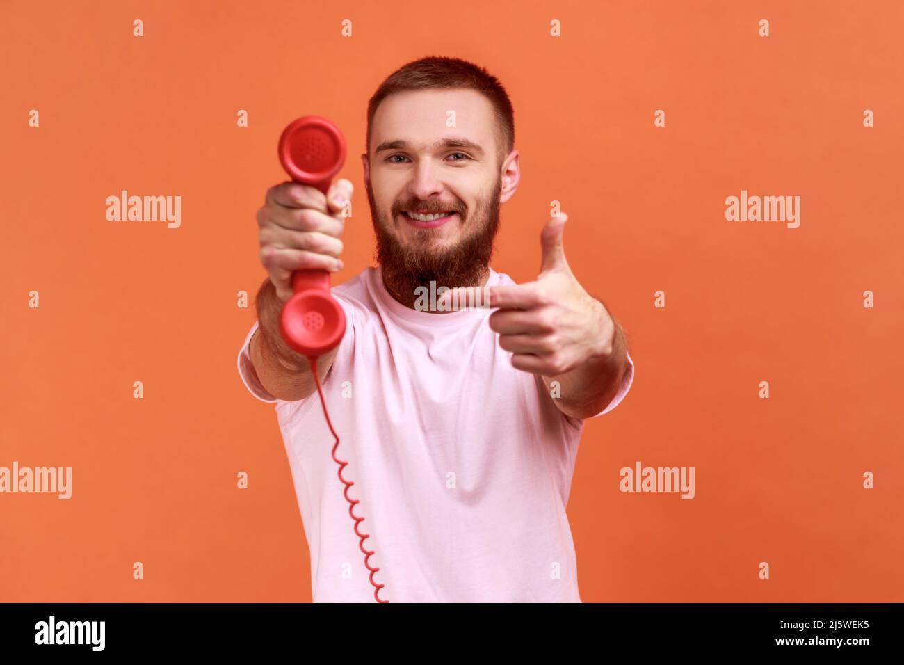 Ritratto di sorridente uomo bearded puntando il dito contro il telefono retro ricevitore alla fotocamera, chiedendo di rispondere telefono, indossando una T-shirt rosa. Studio interno girato isolato su sfondo arancione. Foto Stock