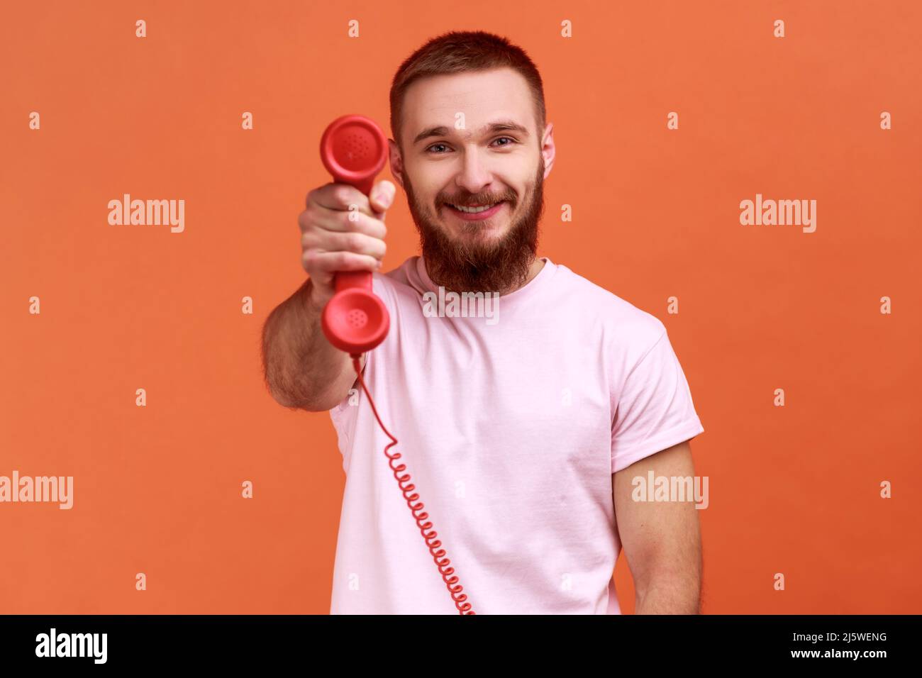 Ritratto di sorridente uomo bearded che tiene e mostra retro telefono cellulare alla fotocamera, chiedendo di rispondere al telefono, indossando T-shirt rosa. Studio interno girato isolato su sfondo arancione. Foto Stock