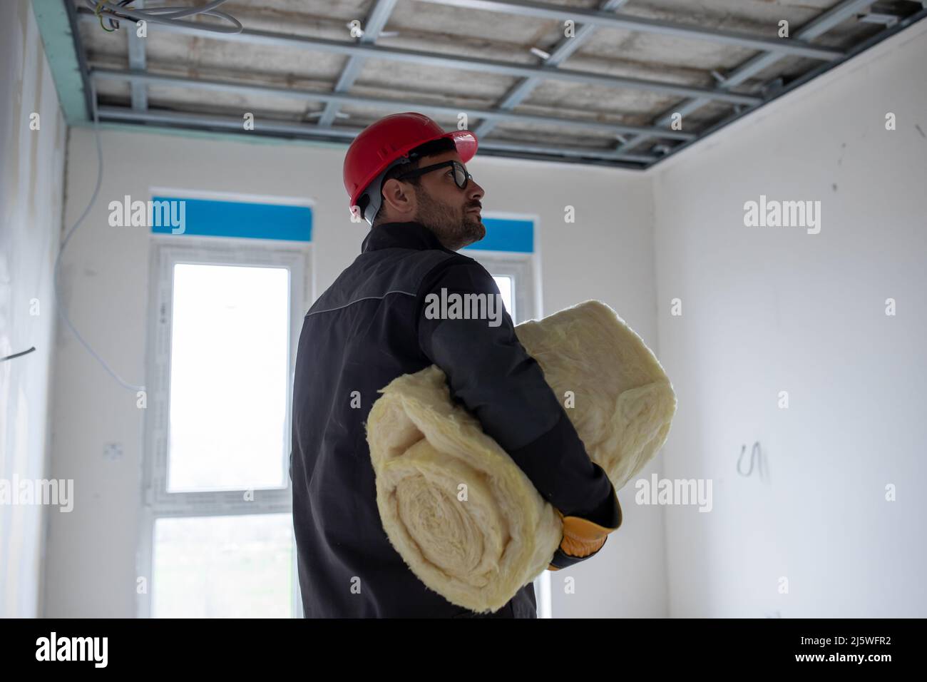 Lavoratore maschio con casco che tiene un rotolo di lana di vetro sotto le sue braccia per l'isolamento in controsoffitto. Foto Stock