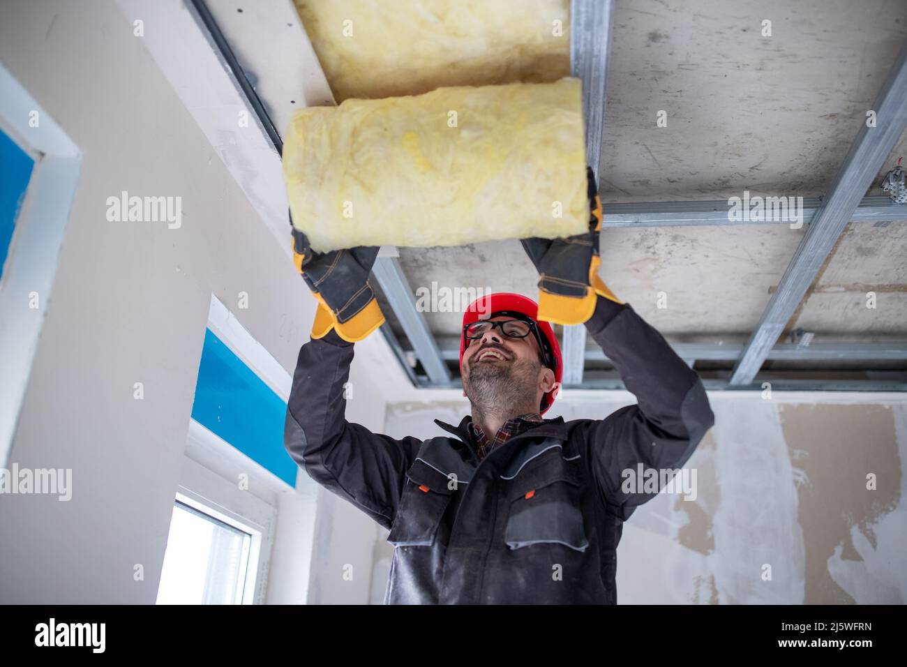 Lavoratore maschile in uniforme con casco installazione lana di vetro per l'isolamento su soffitta. Foto Stock