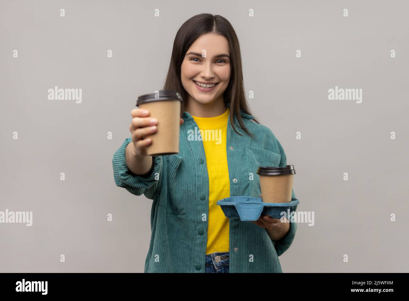 Ritratto di giovane donna felice amichevole che offre caffè, bevande in tazze usa e getta e sorridente, indossando una giacca casual stile. Studio interno girato isolato su sfondo grigio. Foto Stock