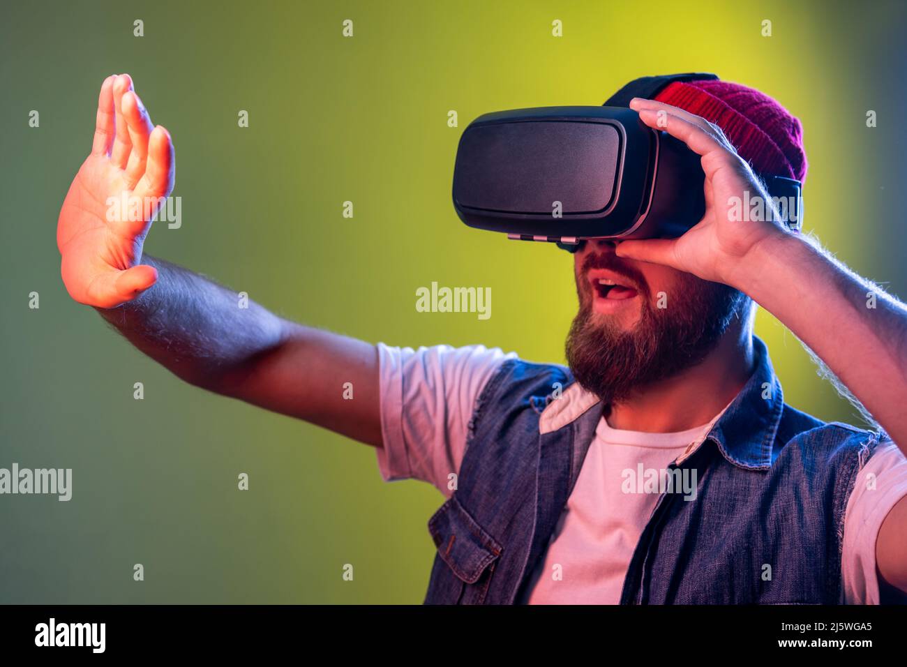 Ritratto di un uomo spaventoso del videogamer con la barba in occhiali vr, allungando il braccio in avanti durante il gioco di realtà virtuale, volto spaventato. Studio interno girato isolato su colorato sfondo di luce al neon Foto Stock