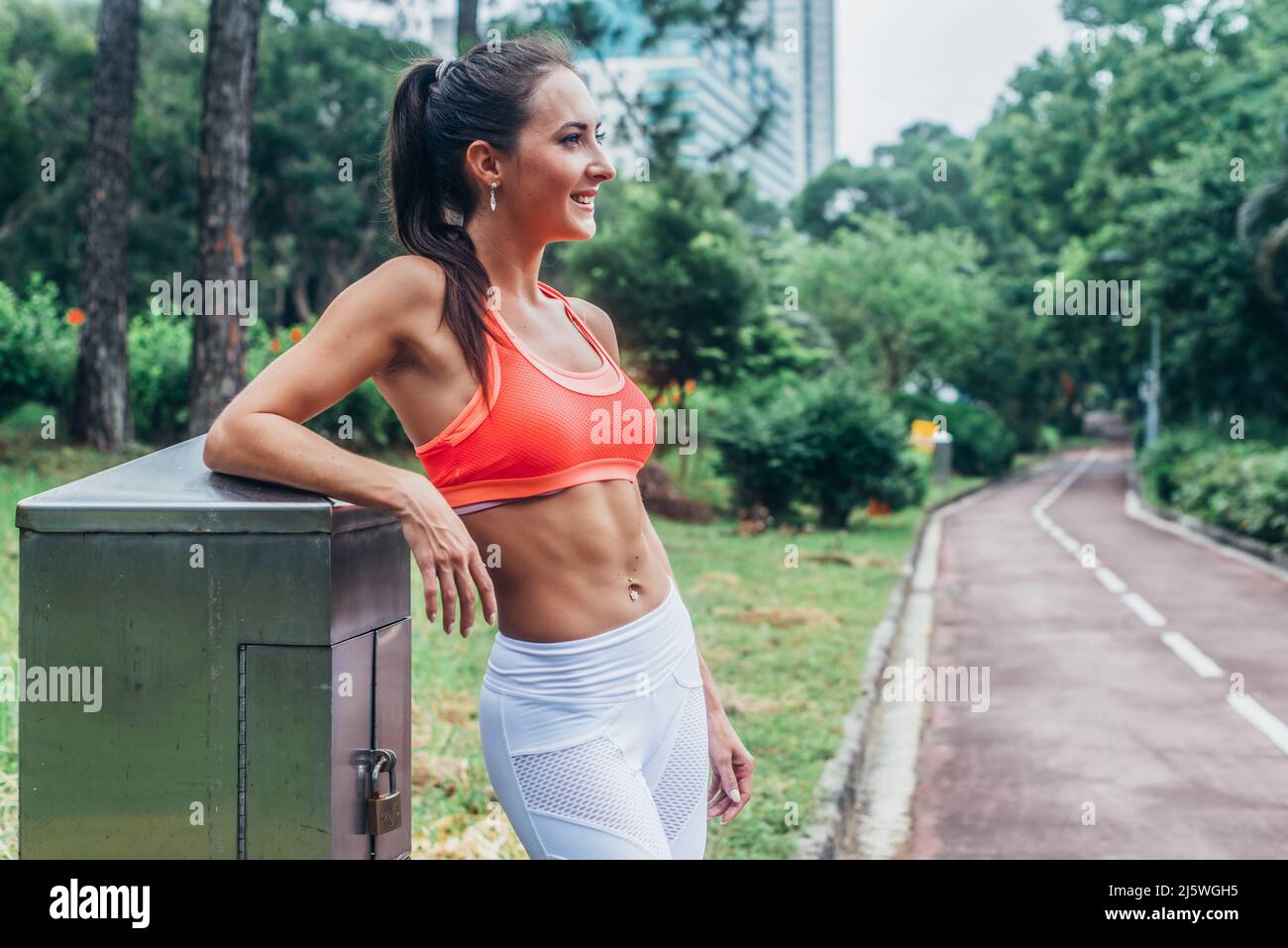 Sottile donna con bruna fitness con sei ABS che indossa un reggiseno sportivo rosa in piedi nel parco della città rilassante dopo l'allenamento guardando lontano dalla fotocamera. Foto Stock