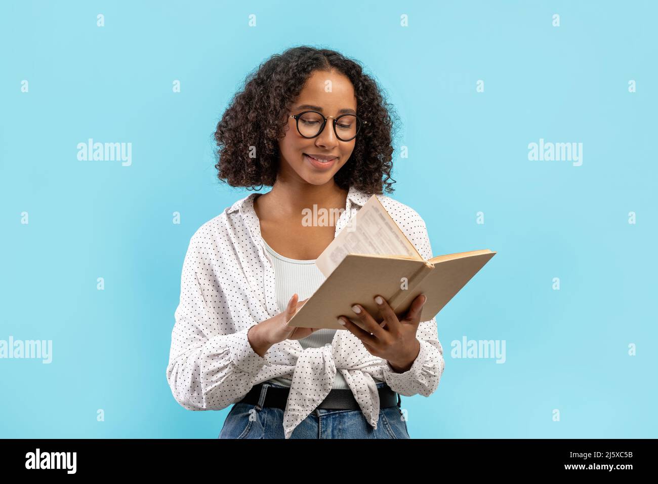 Felice studentessa nera che legge il libro di testo, facendo l'assegnazione domestica, studiando per l'esame su sfondo blu dello studio Foto Stock