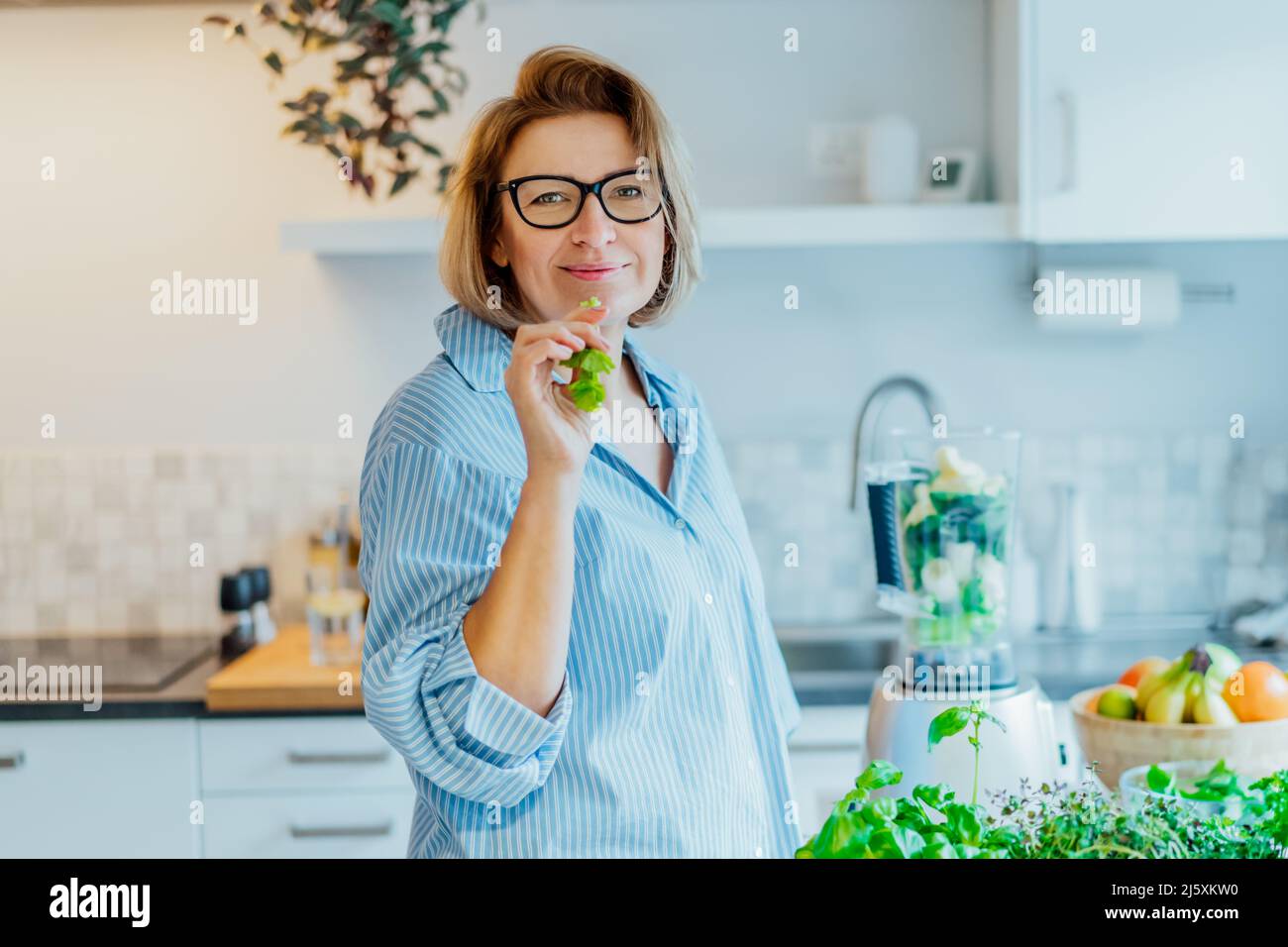 Dieta vegana disintossicante sana. Ritratto di donna di mezza età che mangia un bastone di sedano verde mentre fa frullato in cucina. Sano stile di vita dietetico, noi Foto Stock