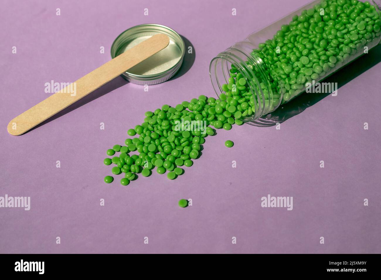 Splendidi granuli di cera verde per la depilazione vengono versati da un vaso trasparente su sfondo viola chiaro, una spatola di legno. Epilazione, depilazione, r Foto Stock