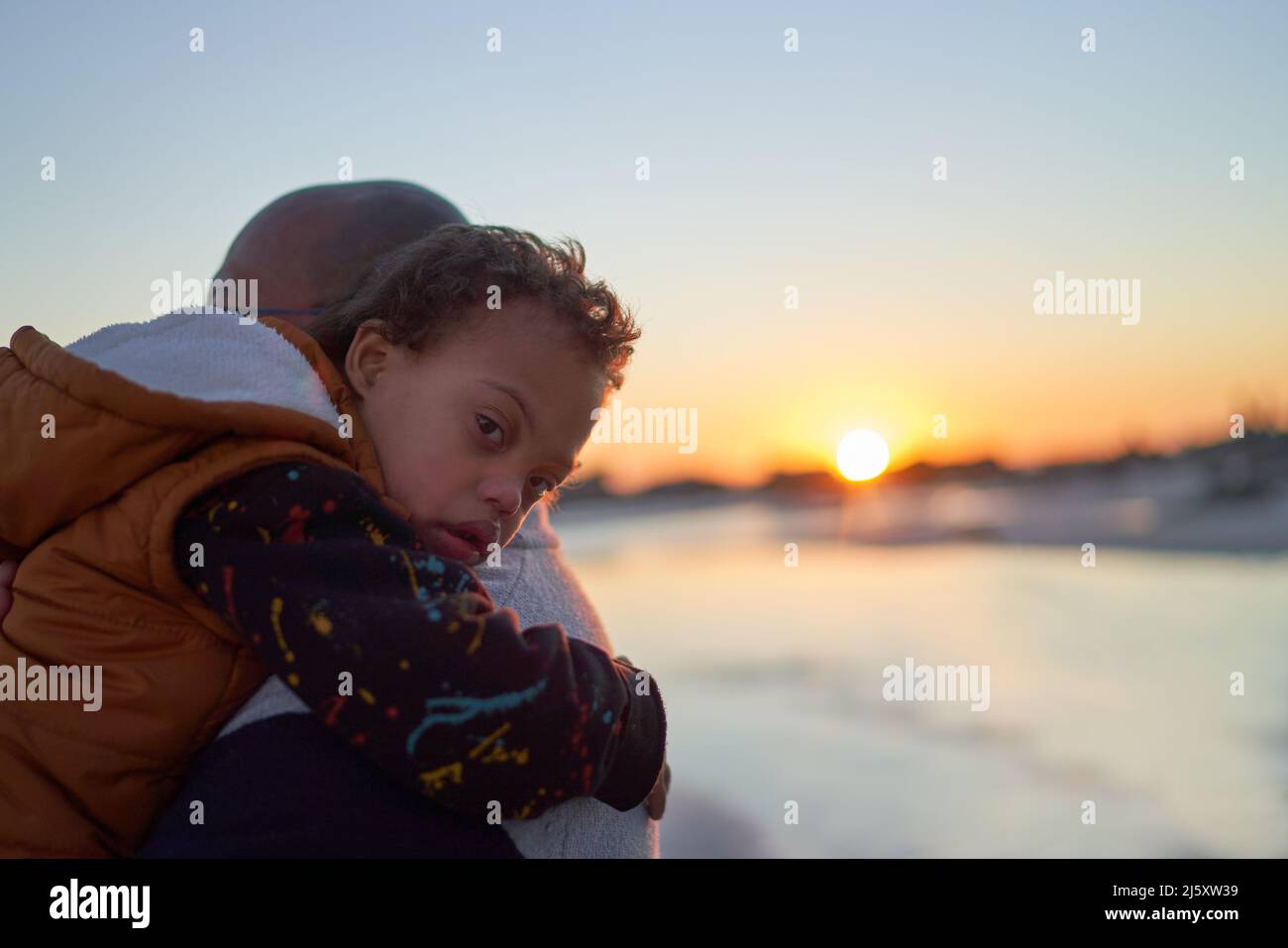 Ritratto ragazzo con sindrome di Down sulla spalla del padre sulla spiaggia al tramonto Foto Stock
