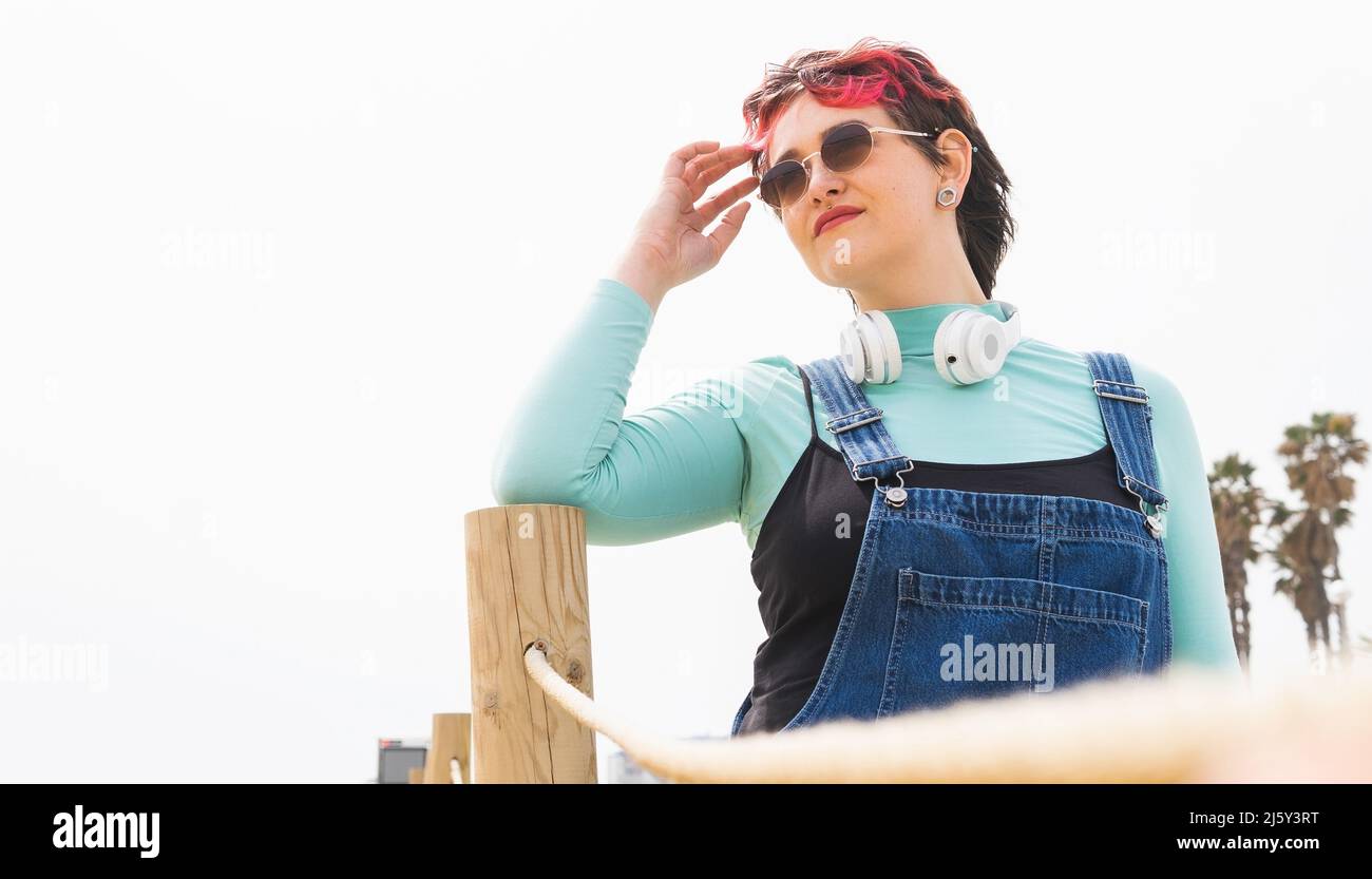 Donna con capelli corti colorati in occhiali da sole e cuffie sul collo guardando lontano mentre tocca la fronte vicino ringhiera di legno Foto Stock