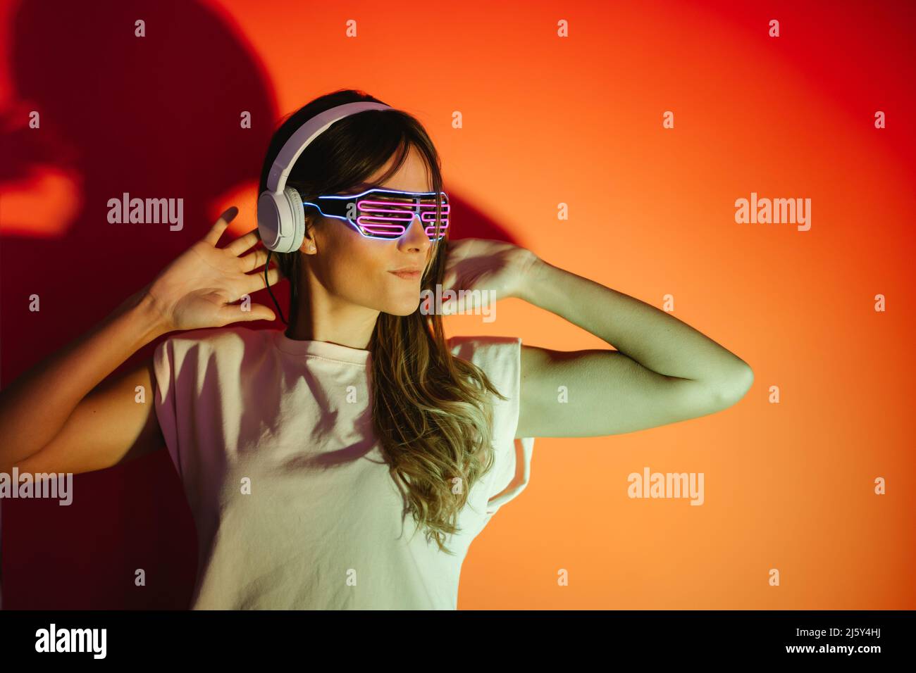 Attraente donna in eleganti tonalità che ascolta la musica con le cuffie wireless mentre si alza su sfondo rosso con ombra in uno studio moderno Foto Stock