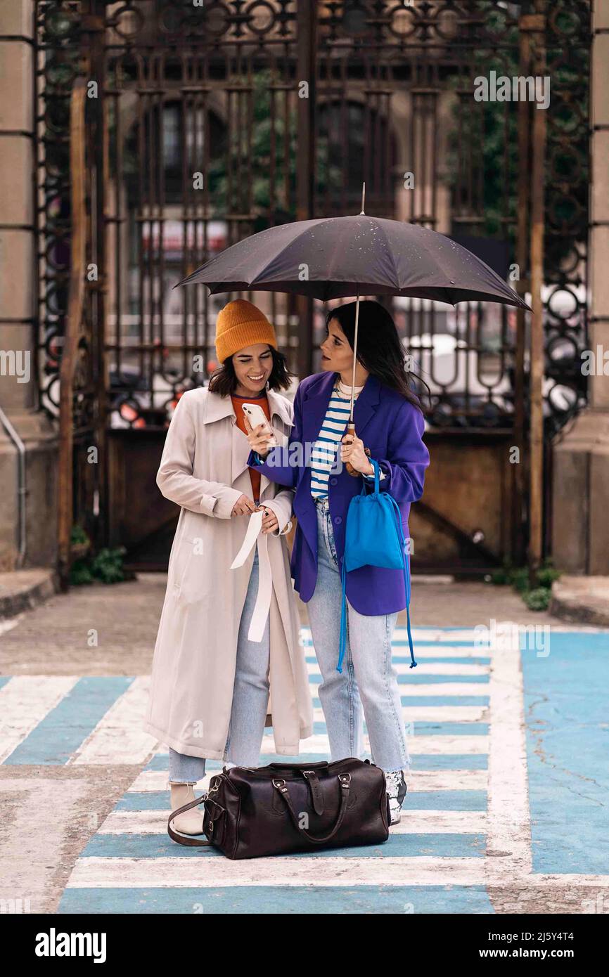 Corpo pieno di amici positivi femminili con ombrello surf social media sul cellulare, mentre in piedi insieme sulla strada in tempo piovoso Foto Stock