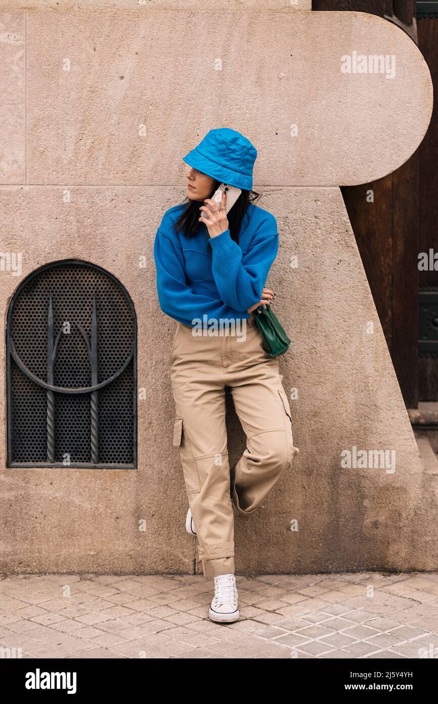 Corpo completo di donna in abbigliamento casual e cappello che hanno conversazione telefonica mentre si sta in piedi su un passaggio pavimentato vicino all'edificio in città Foto Stock