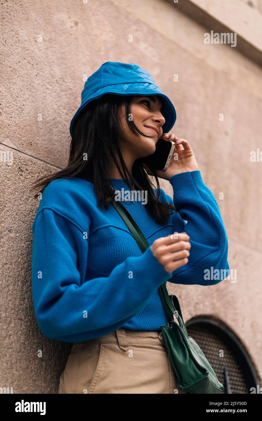 Da sotto femmina in abbigliamento casual e cappello che hanno conversazione telefonica mentre si sta in piedi su strada asfaltata vicino edificio in città Foto Stock
