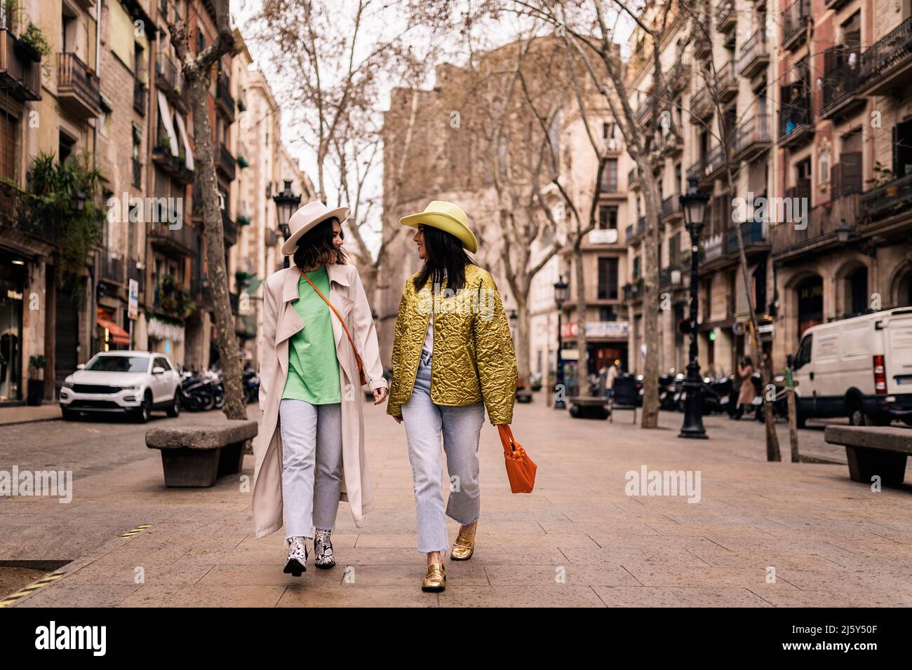 Corpo completo di amici femminili in cappelli eleganti che si guardano l'un l'altro mentre passeggi lungo gli edifici su un passaggio pavimentato in città Foto Stock