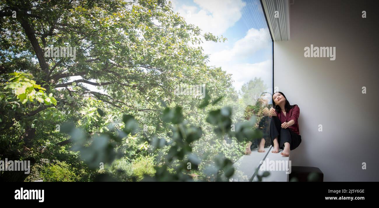 Giovane donna pensierosa seduta alla finestra con vista sugli alberi Foto Stock