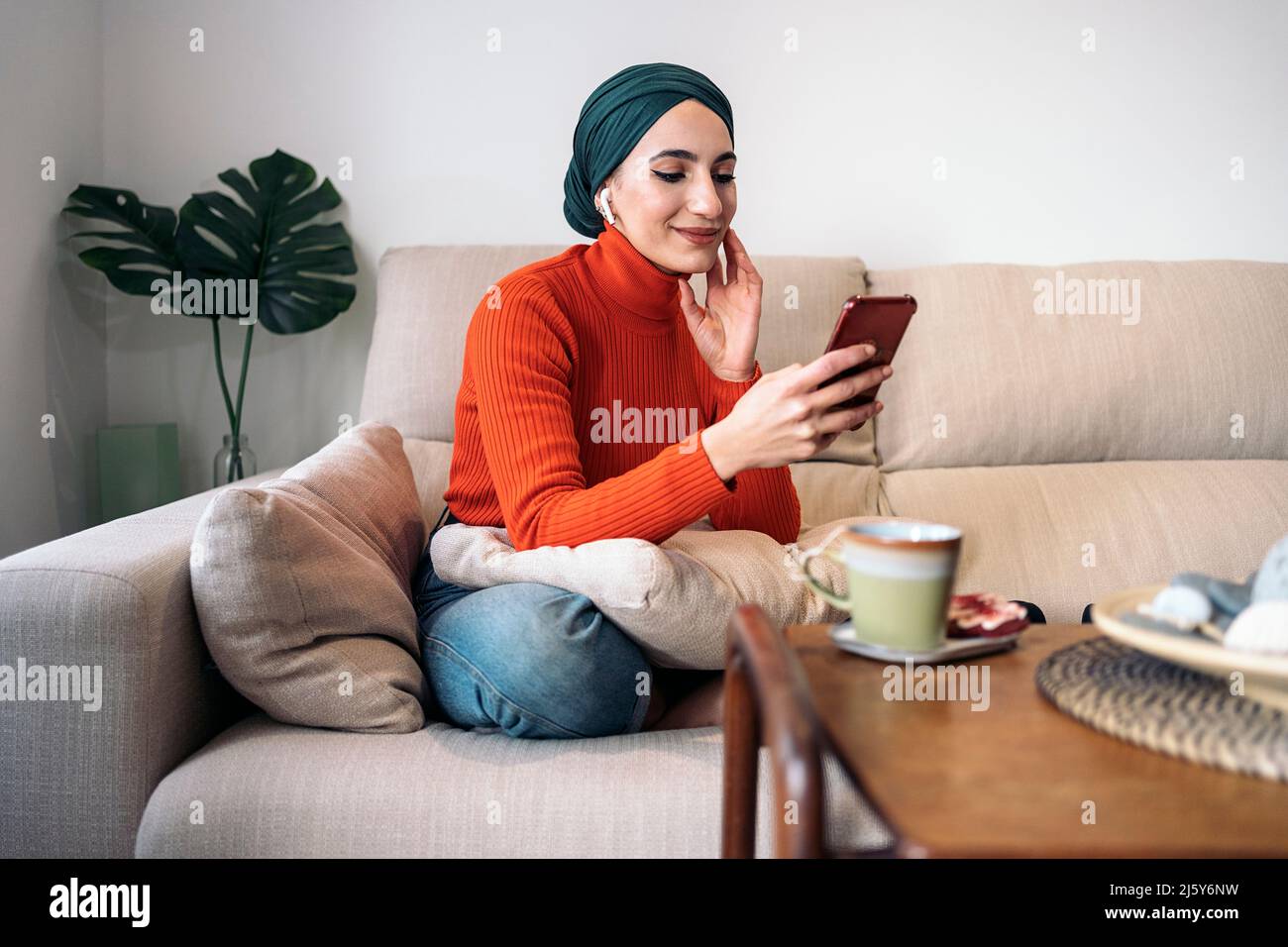Donna musulmana positiva in abiti casual e velo toccare il viso e navigare sui social media sul cellulare mentre si siede a croce sul divano e lis Foto Stock
