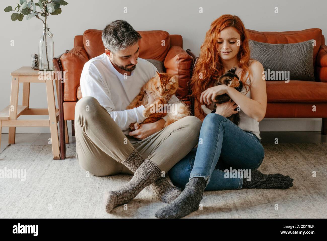 Coppia carezzing carino gatti colorati in mani mentre si siede insieme sul pavimento vicino poltrona in soggiorno chiaro a casa Foto Stock