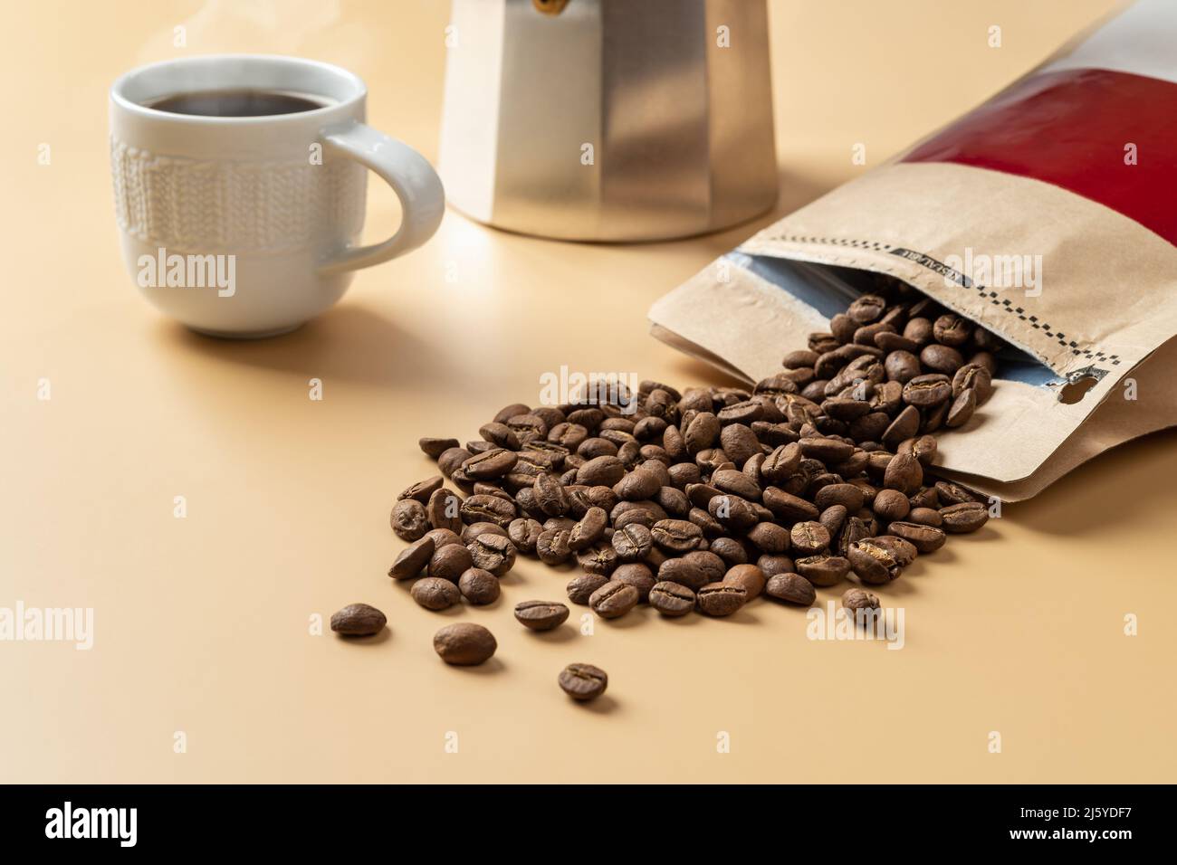 I chicchi di caffè organico fuoriescono da un sacchetto di carta marrone su sfondo beige. Chicchi di caffè torrefatti interi, una tazza di caffè caldo e una macchina per il caffè con geyser. Foto Stock