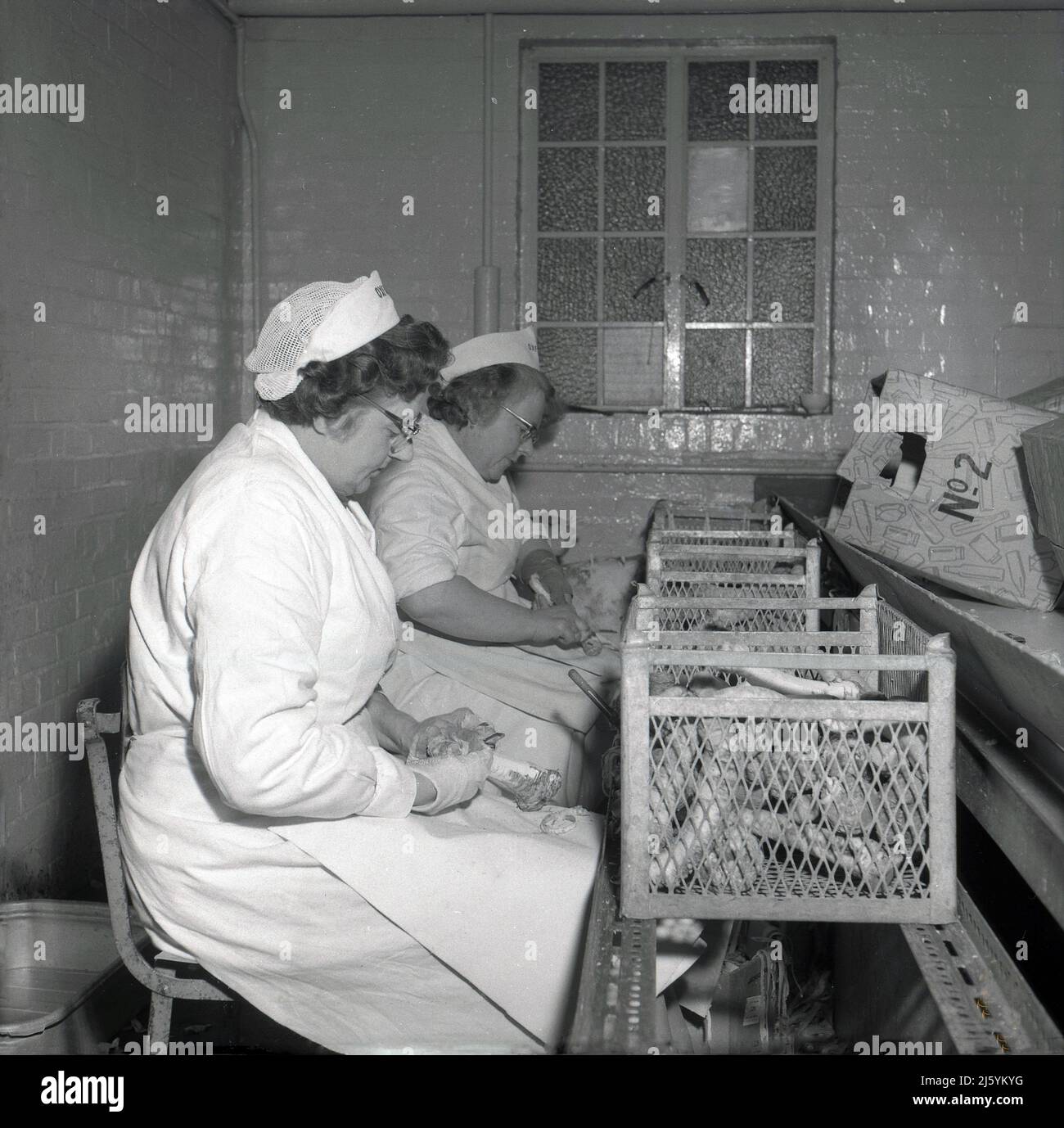 1960s, storica, due lavoratrici che indossano cappotti bianchi e retine che si siedono pelando le verdure ad una società locale di conservazione della frutta, Oxford, Inghilterra, Regno Unito. Foto Stock