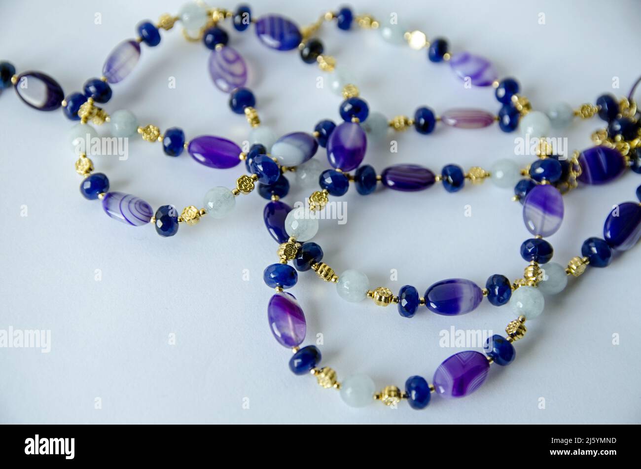 Una collana fatta a mano da perle di acquamarina, agata viola e quarzo blu, intrecciata con perle placcate oro e avvolta su una superficie bianca che lo mostra Foto Stock