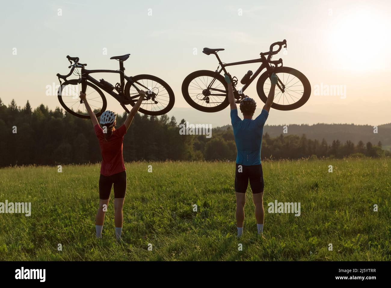 Due silhouette per ciclisti che tengono le biciclette da strada alte sopra le teste, in posa vincente durante il tramonto sul paesaggio di montagna Foto Stock