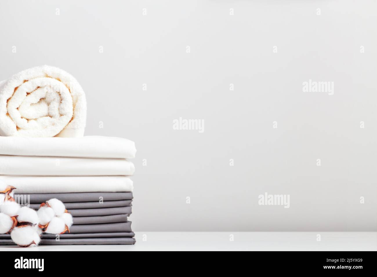 Una pila di lenzuola bianche e grigie, lenzuola e un rotolo di asciugamani su un tavolo con un ramo di cotone. Foto Stock